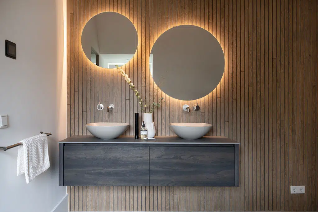 Impressie van een moderne badkamer met grote vloertegels. De badkamer heeft een luxe meubelhoek met donkere wastafel en twee waskommen, designkranen en spiegels met ledverlichting. De meubelwand heeft een houtlook tegel, geplaatst in verticale stroken.