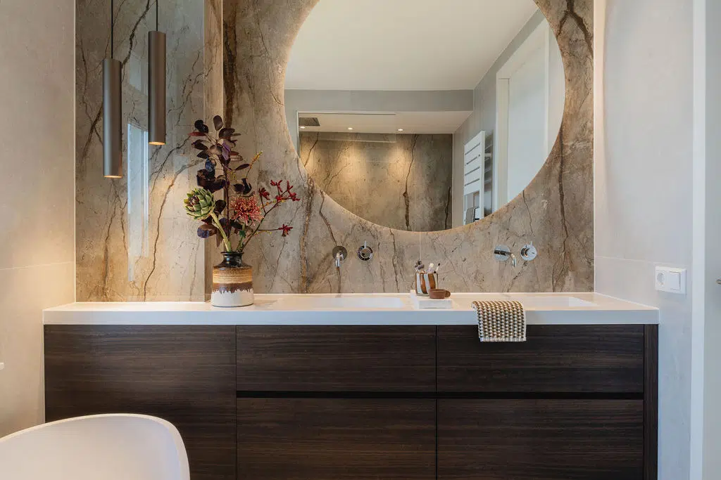 Impressie van een luxe badkamer met natuursteen slabs op de wand. We zien een maatwerk badkamermeubel met luxe ronde spiegel, een wastafel met twee kranen. De kleur chroom van de kranen zorgt voor een fris detail. Op deel van de wastafel is ruimte voor decoratie. We zien een vaas met aardse schildering en fraaie planten.