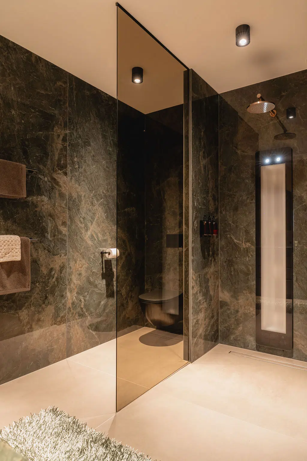 Impressie van een design badkamer, voorzien van een inloopdouche met Sunshower en Gessi kranen, en een toiletruimte. De ruimtes zijn gescheiden door een glazen douchewand, en hebben beiden groene tegels in een marmerlook.