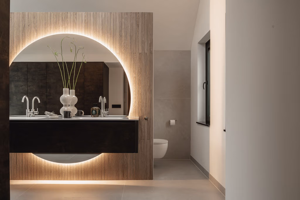 Binnenkomst bij een moderne badkamer met donker badkamermeubel, bevestigd aan een wand met houtlook tegels. Achter het meubel loopt een royale ronde spiegel met verlichting. De spiegel steekt zowel aan de onderkant als bovenkant van het meubel uit.