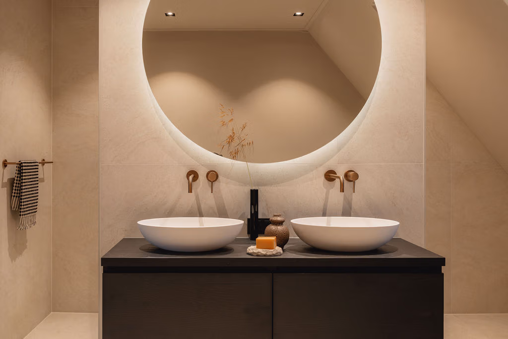 Impressie van een badkamer nieuwbouw in moderne stijl, voorzien van een houten badkamermeubel, twee waskommen en ronde spiegel met verlichting.