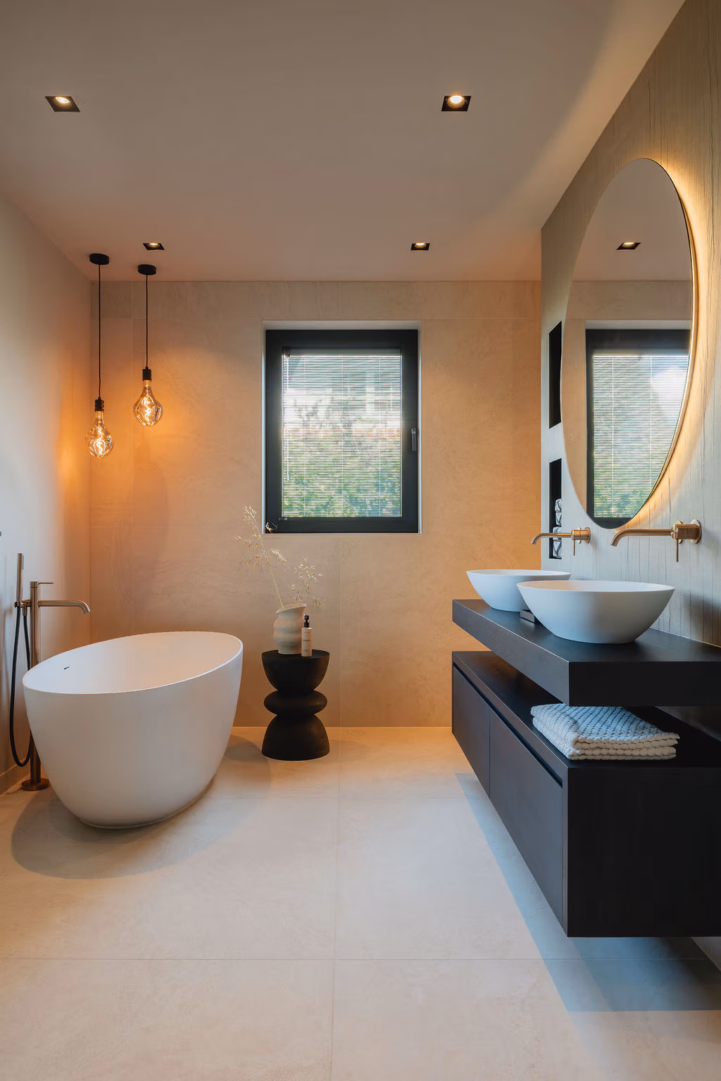 Impressie van een badkamer nieuwbouw in moderne stijl, voorzien van vrijstaand bad, een tegelsoort, houten badkamermeubel, twee waskommen en ronde spiegel met verlichting.