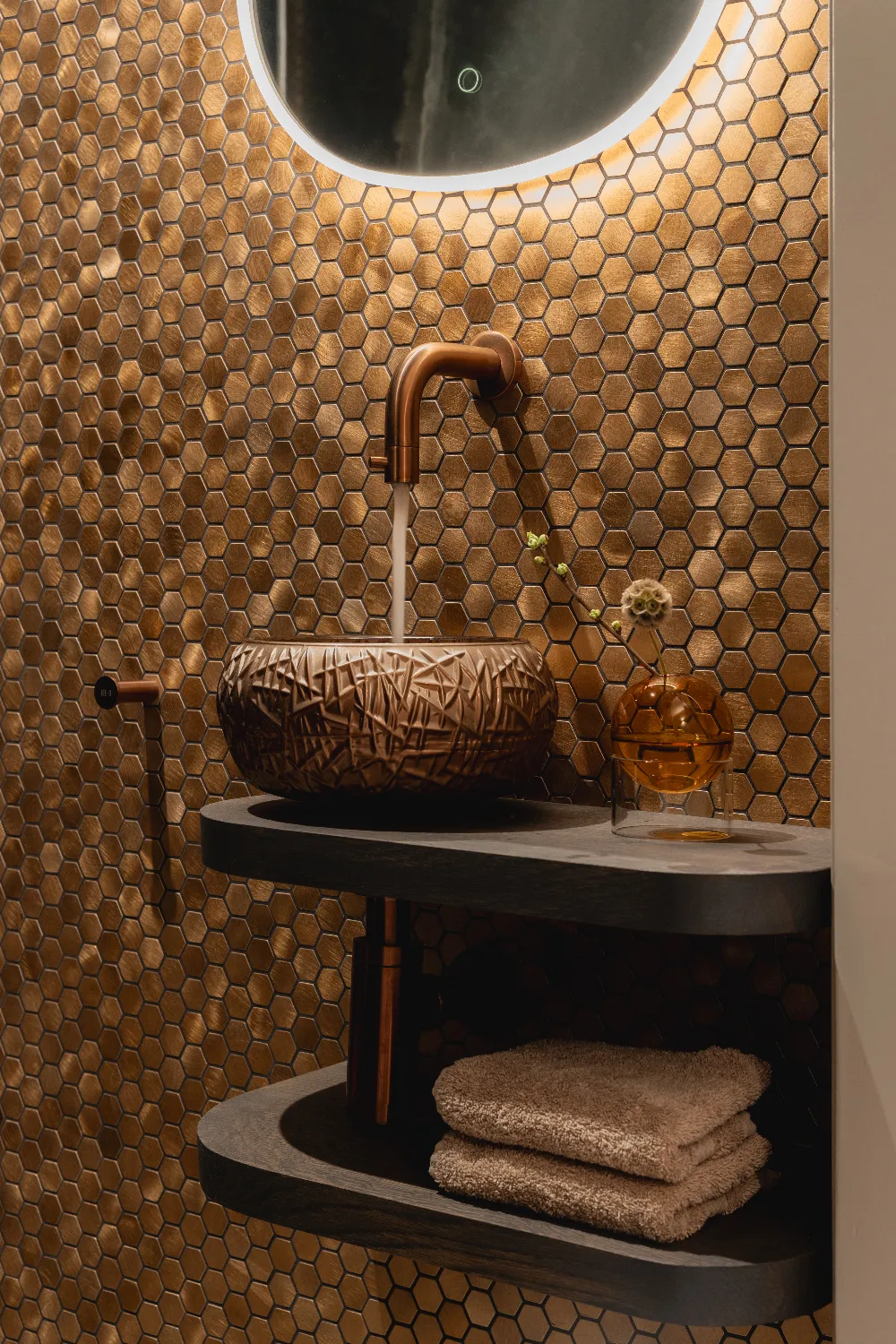 Een close-up van een bronskleurige waskom op een houten meubel, met een muur van metallic hexagon mozaïektegels.