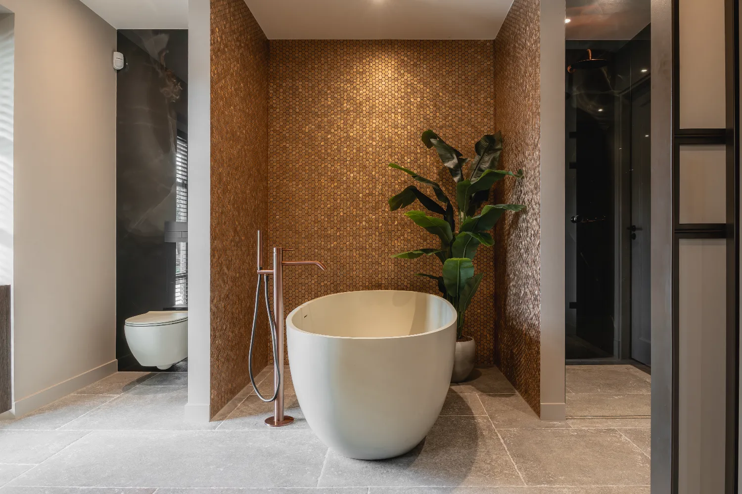 Een badkamernis met een vrijstaand bad, metallic hexagon mozaïektegels, een bronzen kraan en een inloopdouche met zwarte tegels.